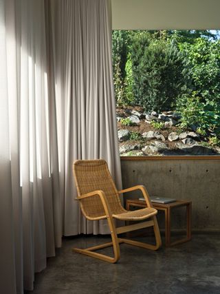 A wooden armchair is set in the corner, next to a large window. A white curtain covers one side of the window.