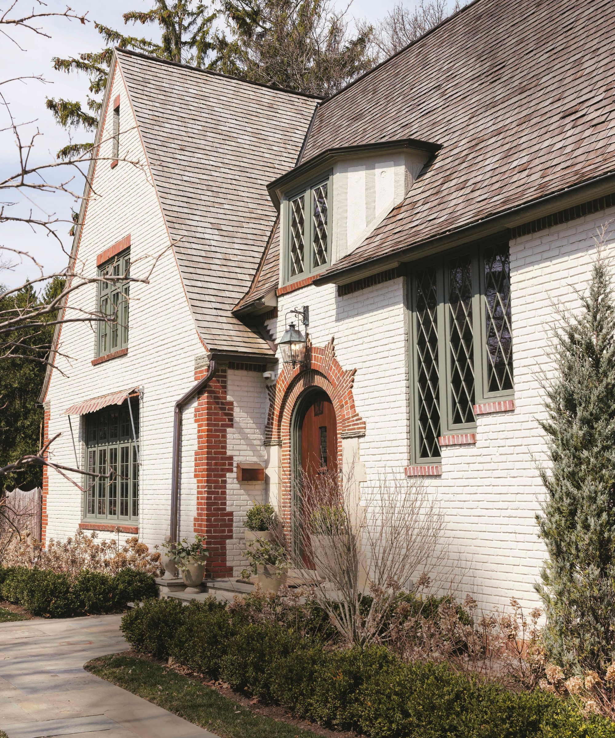 the exterior of a white brick painted tudor home with brick arched doorway, soft greenwindows and hedge planting