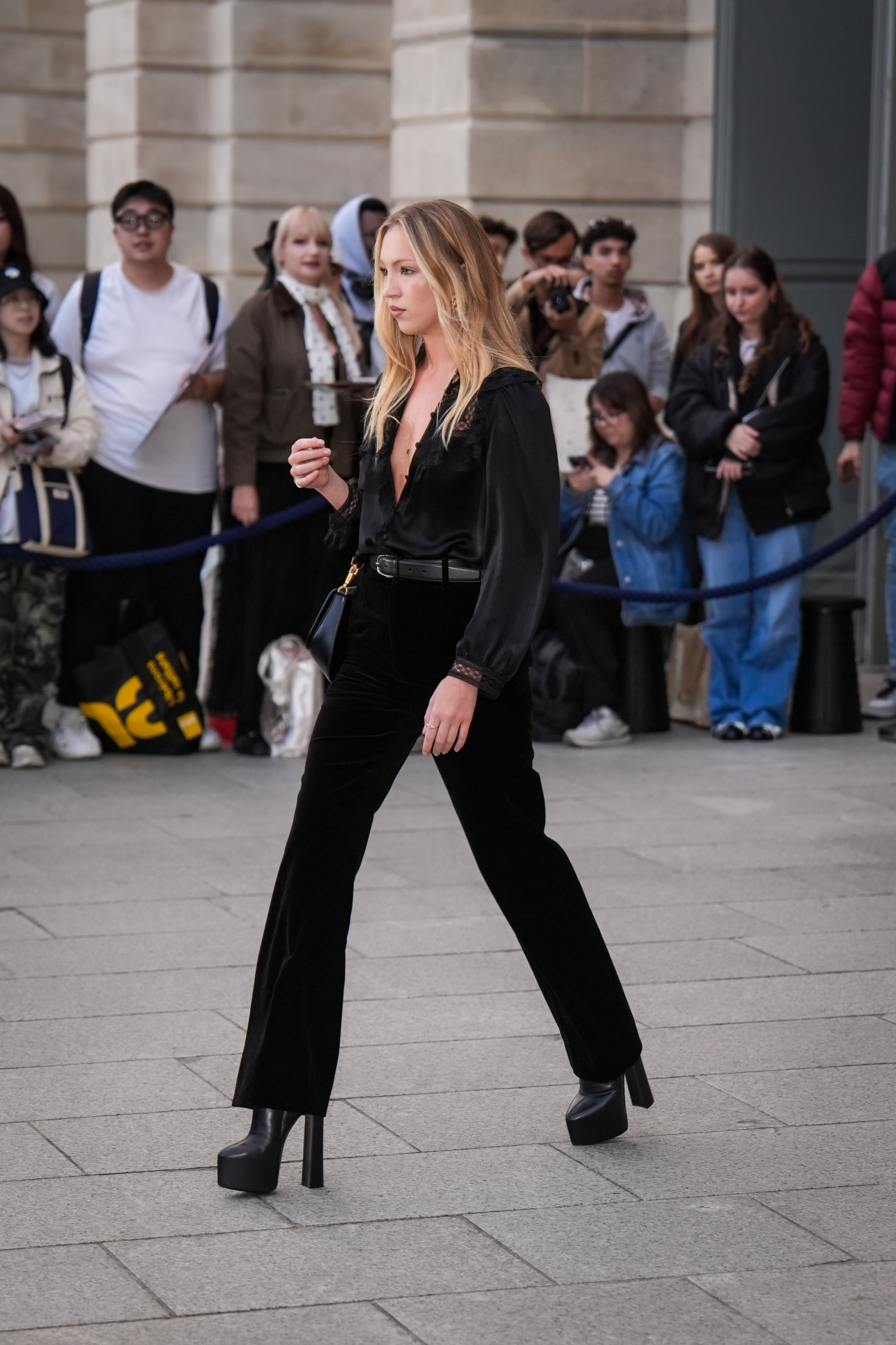 Lila Moss wears black flared jeans with a black blouse and heeled platform boots.