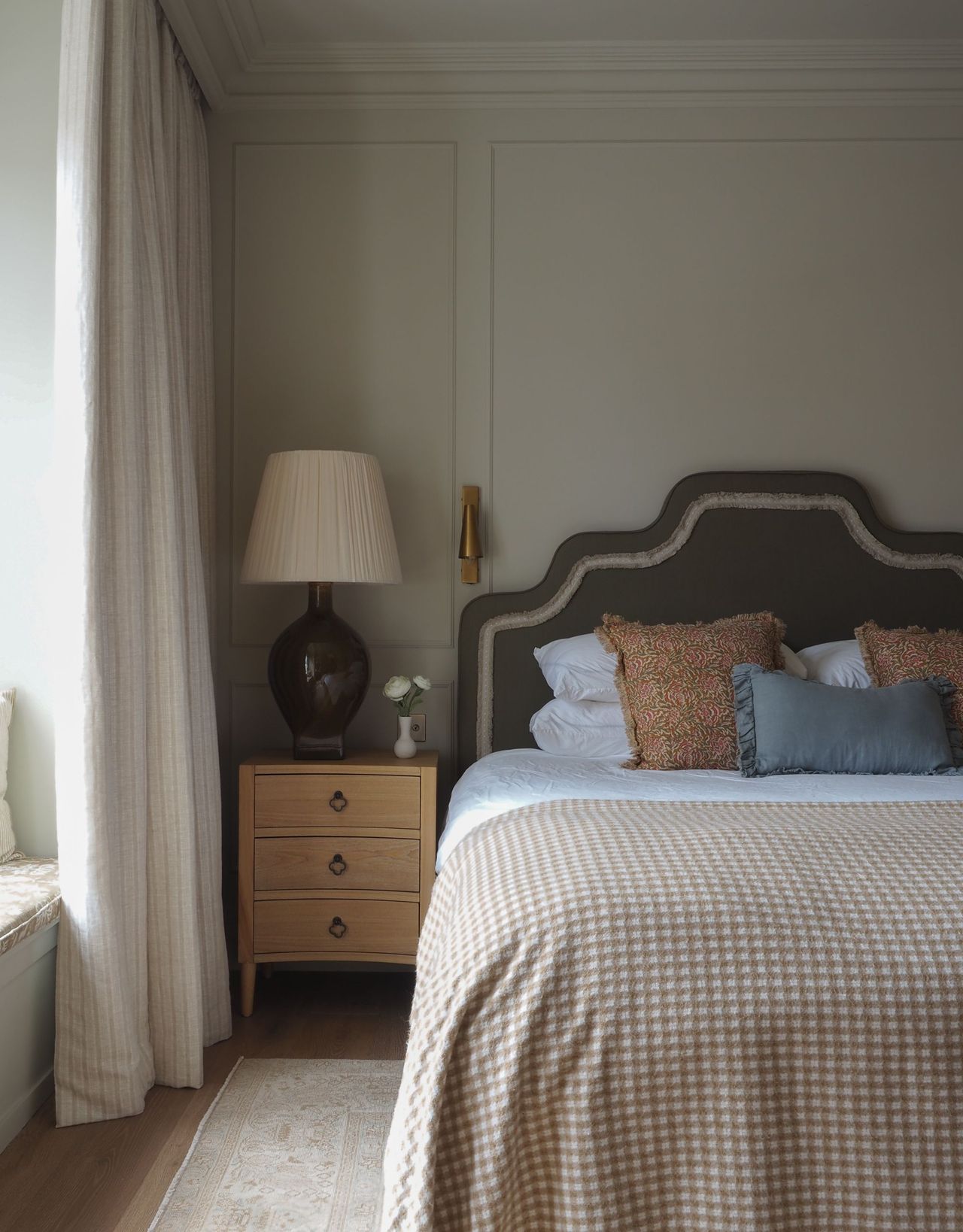 A modern neutral bedroom with a green headboard, white striped curtains, a wooden bedside table with a large lamp on top, and checkered bedding