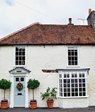 exterior of Georgian cottage with whitewashed brick walls and pale green front door with spring wreath and olive trees on either side and georgian bay window