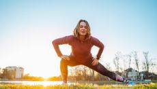 Woman performing side lunge stretch outdoors. There is a lake behind her with a low sun on the horizon. She is wearing sports leggings and a red long-sleeved top. 