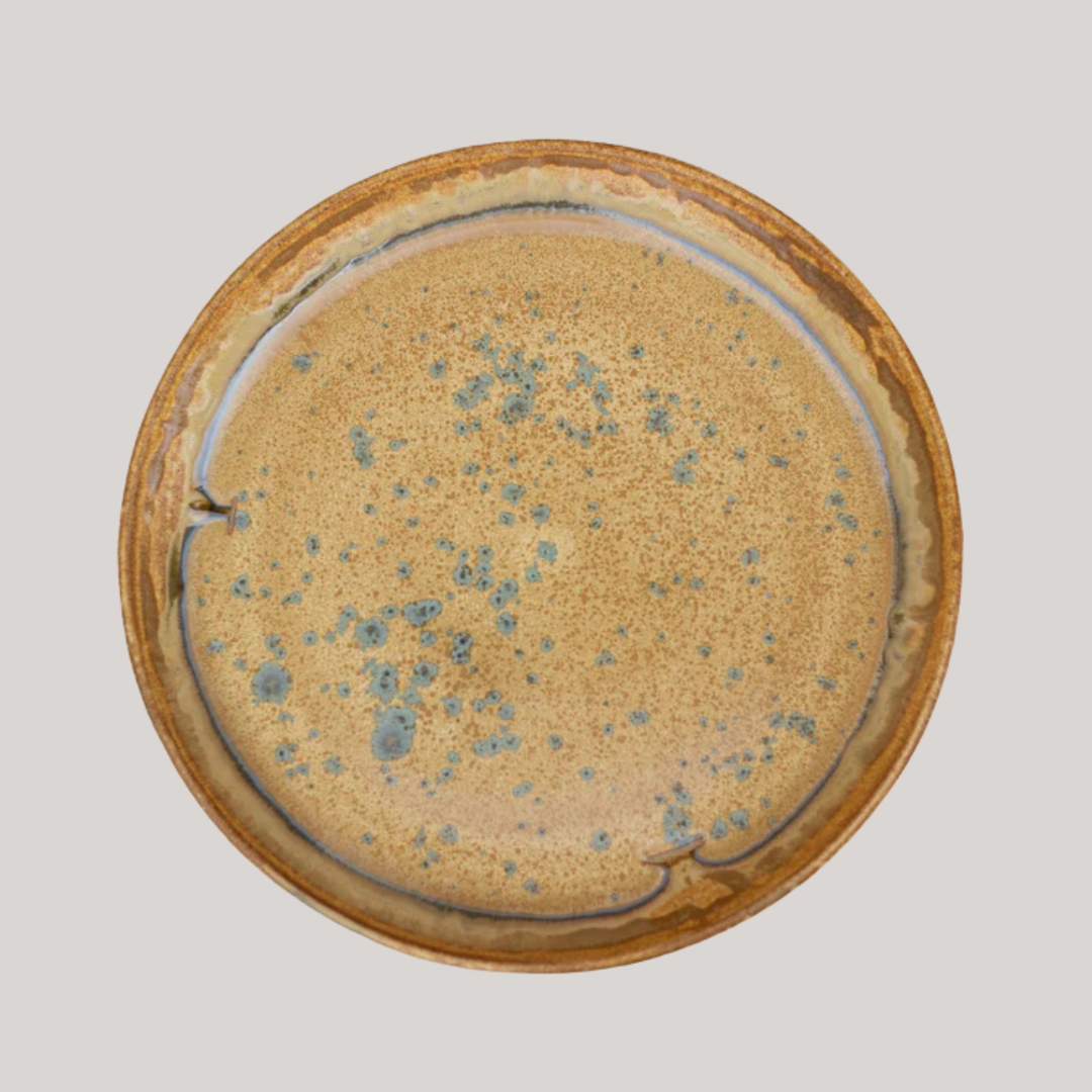 An organic earthenware plate with golden and green glacing sits against a pale brown background.