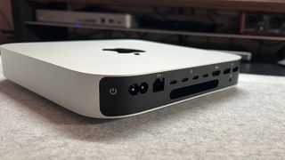 The Mac Mini Pro M2 is mini pc. It's a thin, square shape with rounded corners (20 x 20 x 3.5 cm). It's silver in color and has the Apple logo in the center of it (a black apple with a bite taken out of it). Here we see a close up of one edge – it's black and has several ports/inputs, such as a headphone jack.