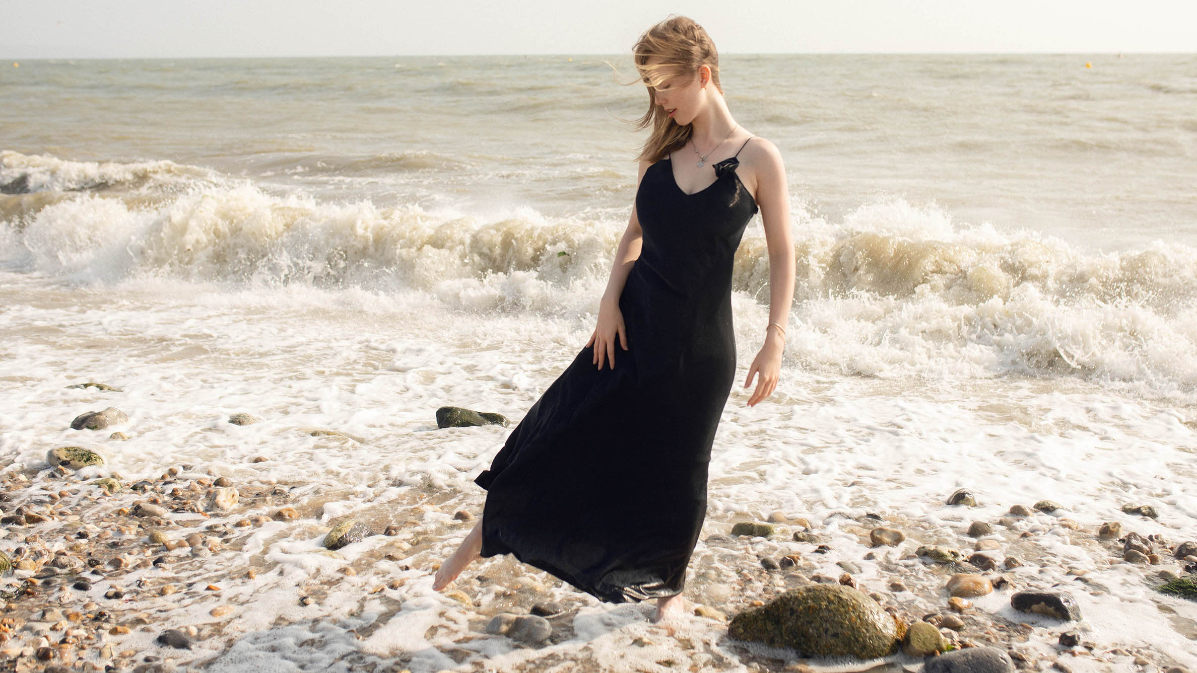 A woman in a black dress and blond hair, standing on a stone beach in front of the water, it is windy and her dress and hair go with the wind