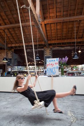 View of a woman in a black dress on a wooden and rope swing. Behind the swing is a bar with shelves of drinks and a sign in front that reads 'Come and play at King's Cross