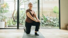 A woman kneels on an exercise mat, with her right knee on the mat and her left foot on the floor in front of her. Behind her are some sliding glass doors, showing a green garden.