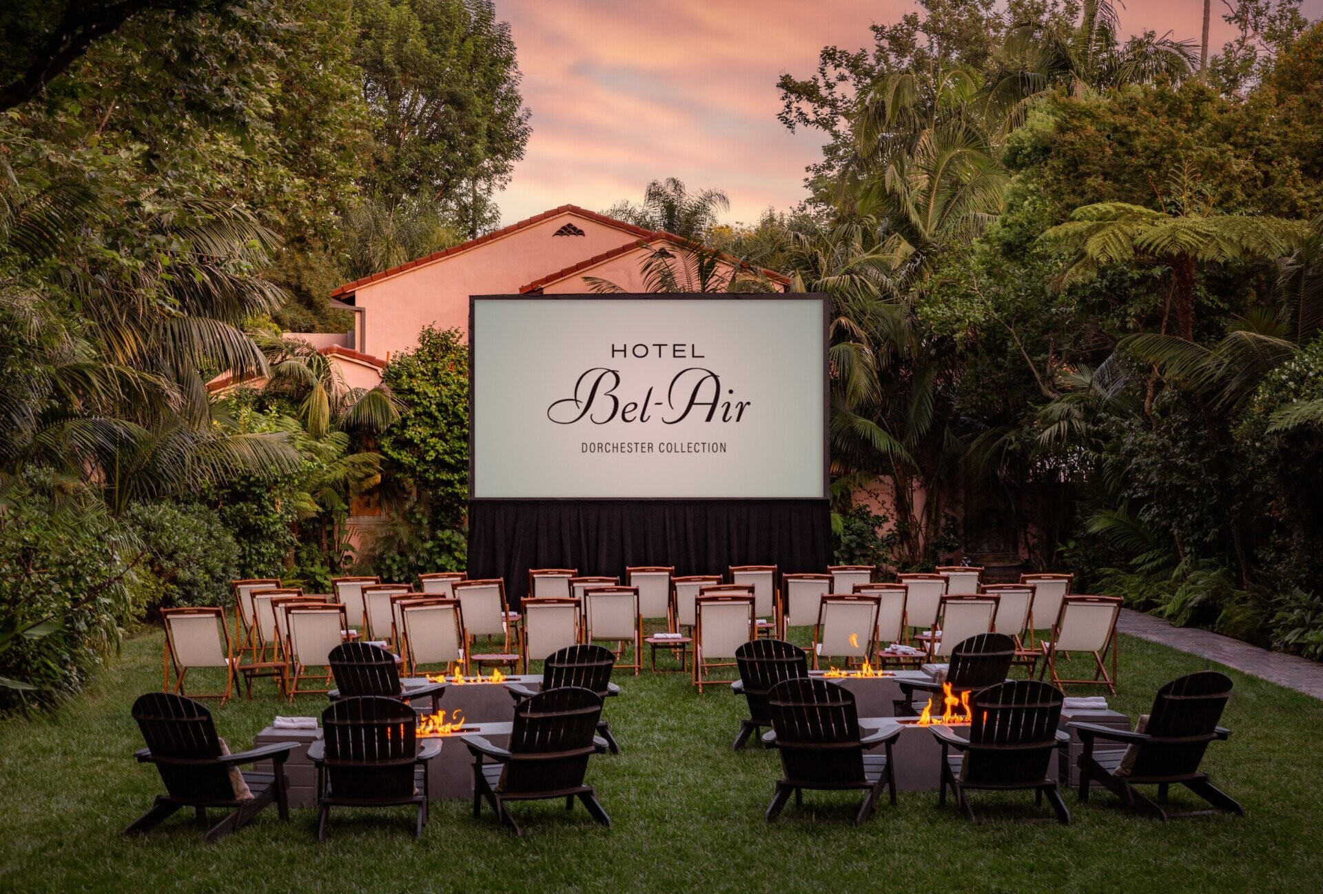 An outdoor cinema setup with white textile and wood deck chairs, as well as black ones, lit-up fireplaces, and lush grass and tropical plants.