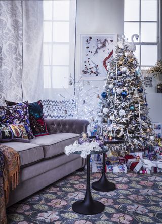 A silver frosted Christmas tree laden with baubles, blue and silver, china ornaments and Christmas tree lights, and sofa and tables.