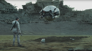 Tom Cruise in the film Oblivion, standing in a ruined sports stadium with a spherical drone aiming its guns at him.