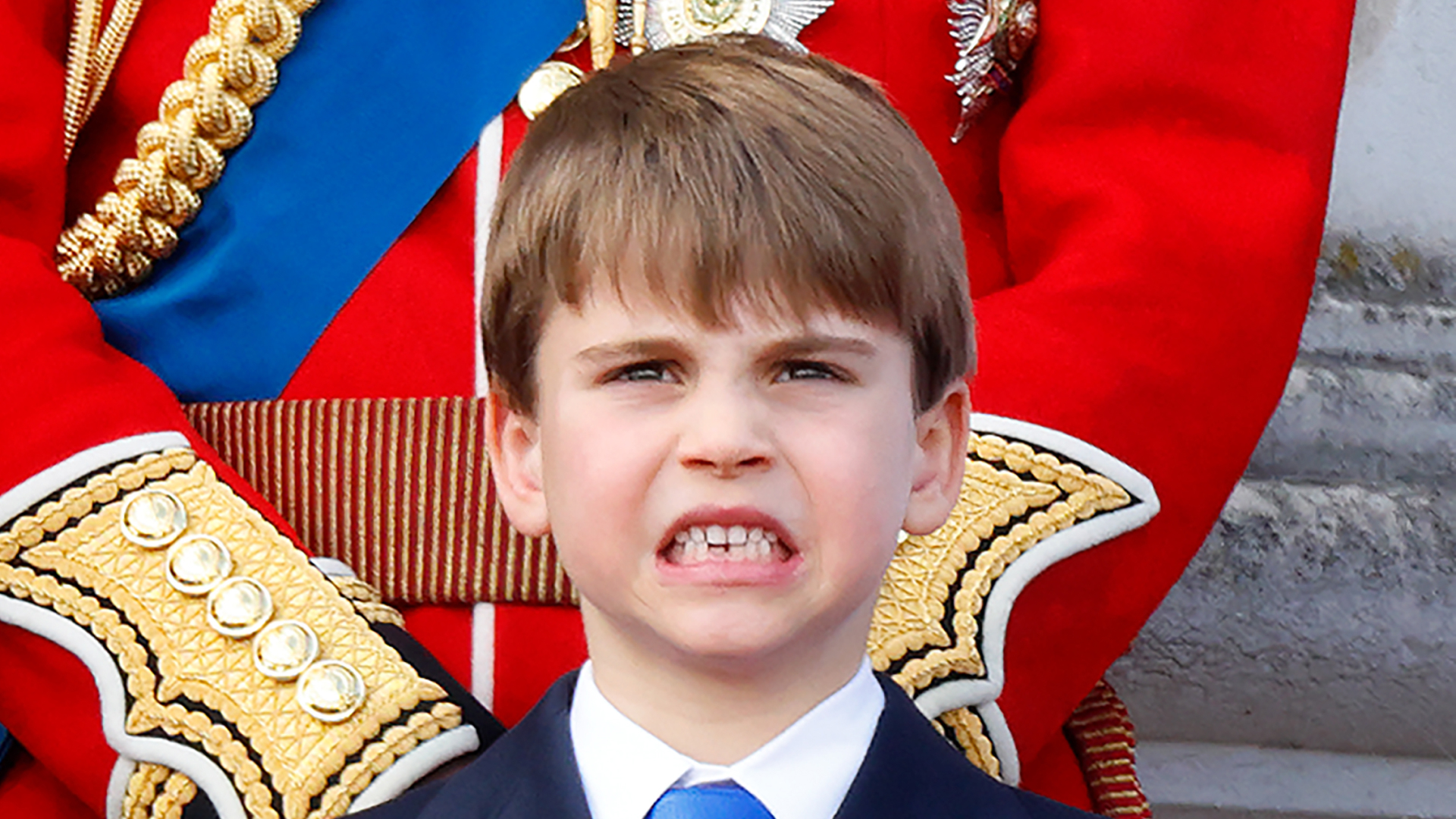 Prince Louis stands on the Buckingham Palace balcony wearing a navy suit with a white shirt and royal blue tie