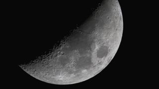 The moon shines with its right half lit and its left veiled in shadow against a black night sky. Dark plains can be seen marking its surface around the equator, while large craters line the line separating night from day. 