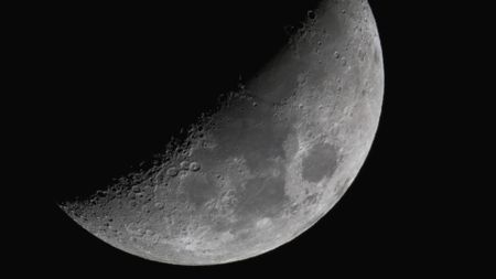 The moon shines with its right half lit and its left veiled in shadow against a black night sky. Dark plains can be seen marking its surface around the equator, while large craters line the line separating night from day. 