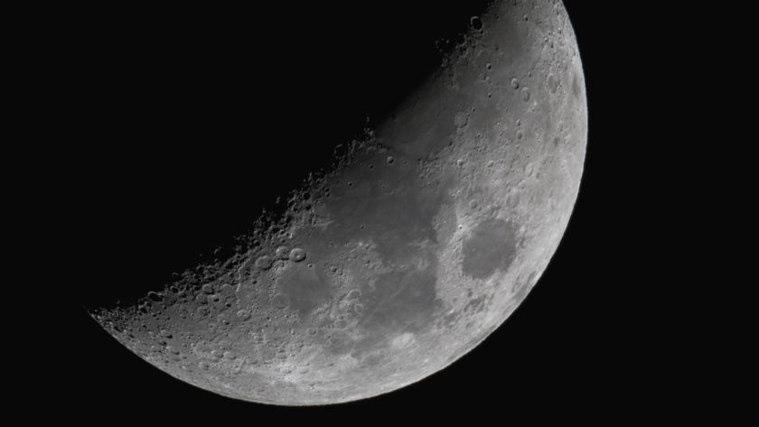The moon shines with its right half lit and its left veiled in shadow against a black night sky. Dark plains can be seen marking its surface around the equator, while large craters line the line separating night from day. 