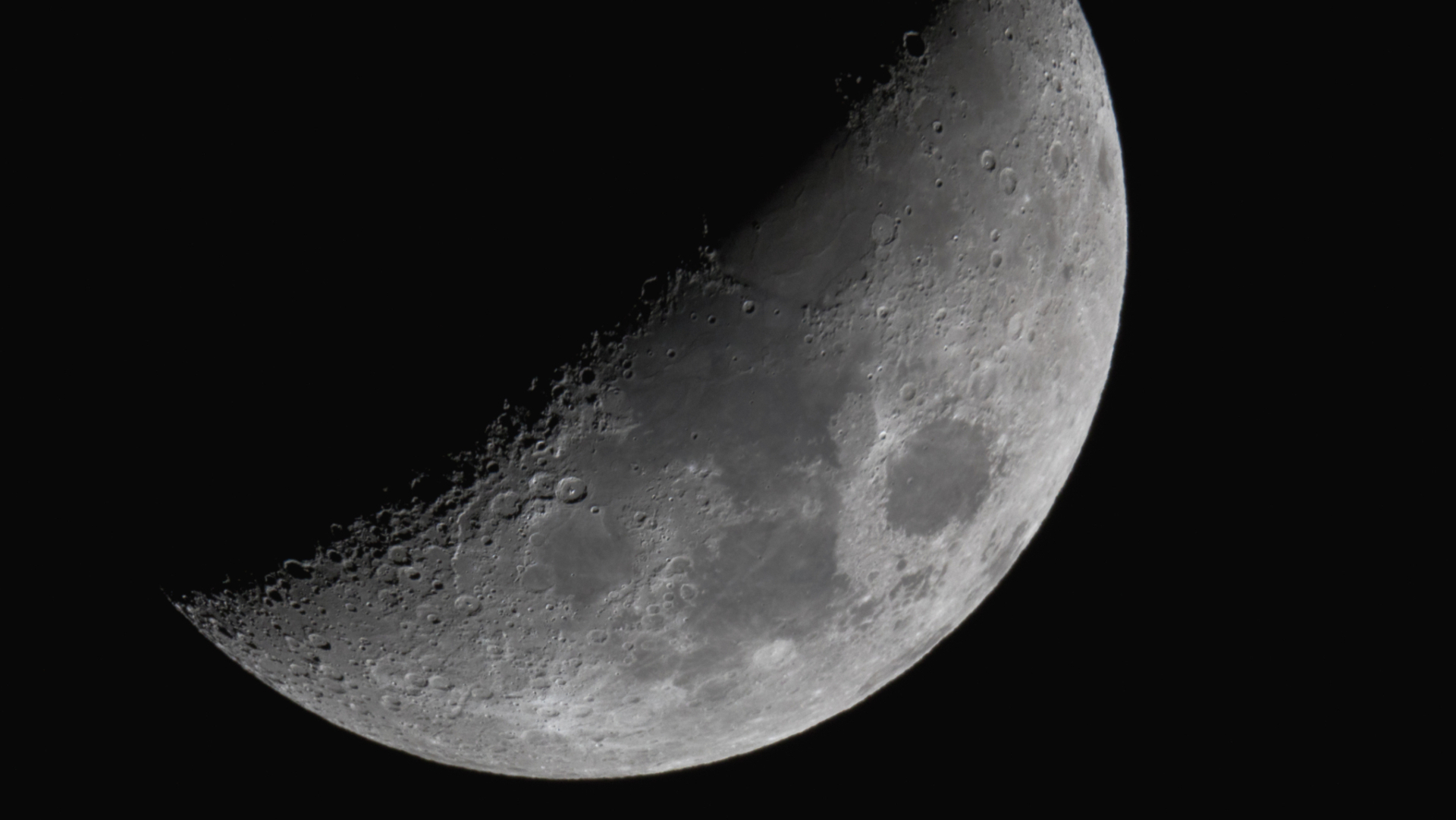 The moon shines with its right half lit and its left veiled in shadow against a black night sky. Dark plains can be seen marking its surface around the equator, while large craters line the line separating night from day. 