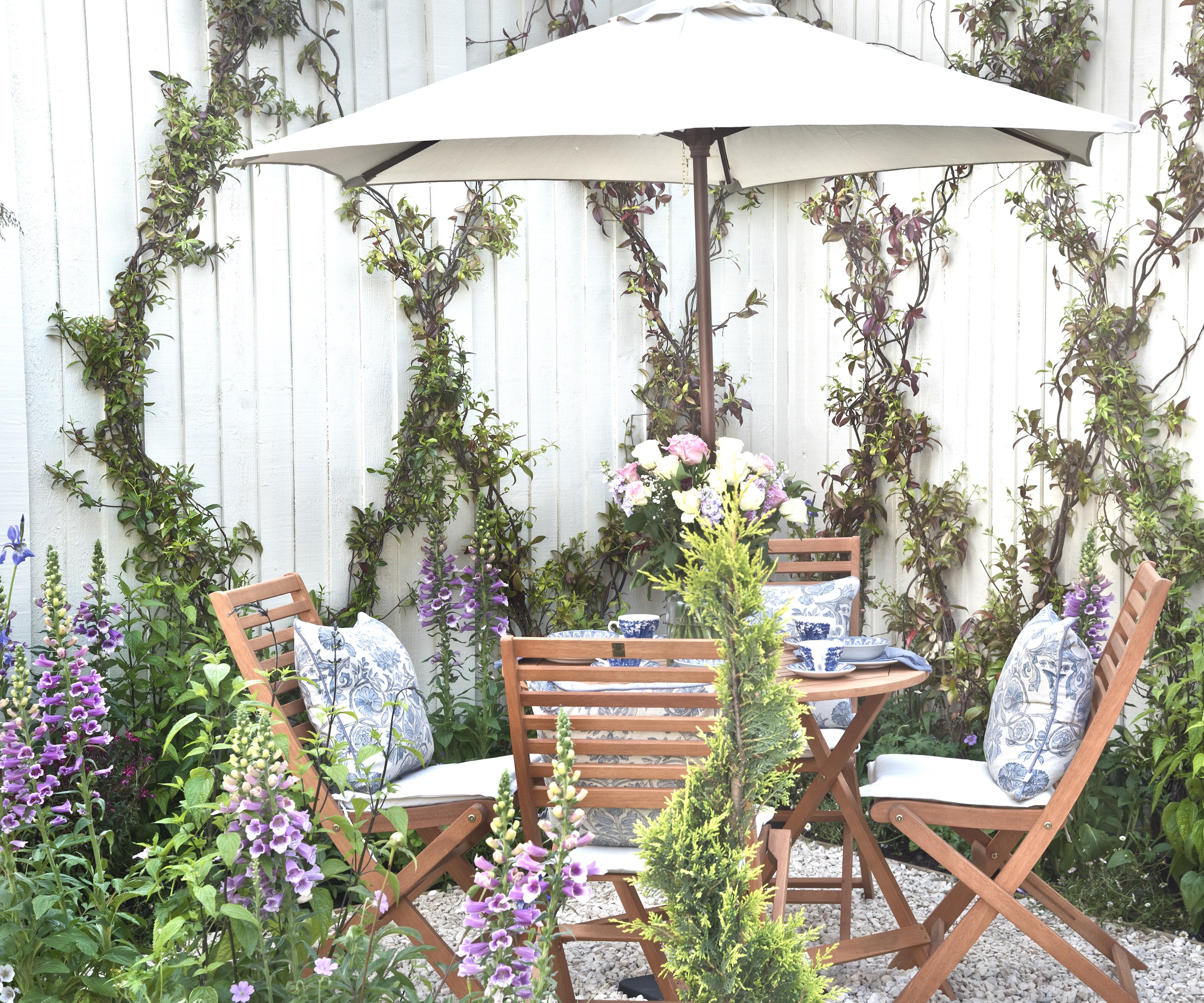 Small round table and chairs in gravel garden, in front of white fence with plants climbing on it and foxgloves in foreground