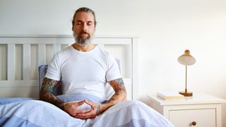 Man with tattoos sits upright in bed practicing a sleep hack to help him fall asleep faster