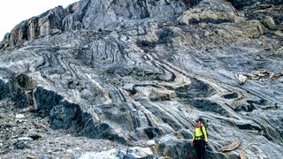 A person with a neon yellow jacket stands in front of a huge wall of rock with cool striations.