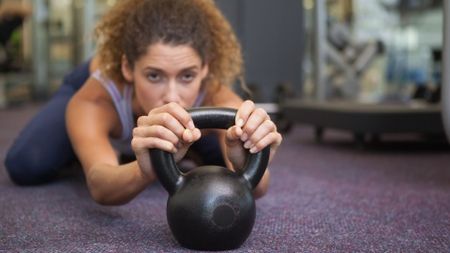 A woman with curly hair lies on her front clutching a cast-iron kettlebell. She is wearing blue leggings and a mauve sports bra.