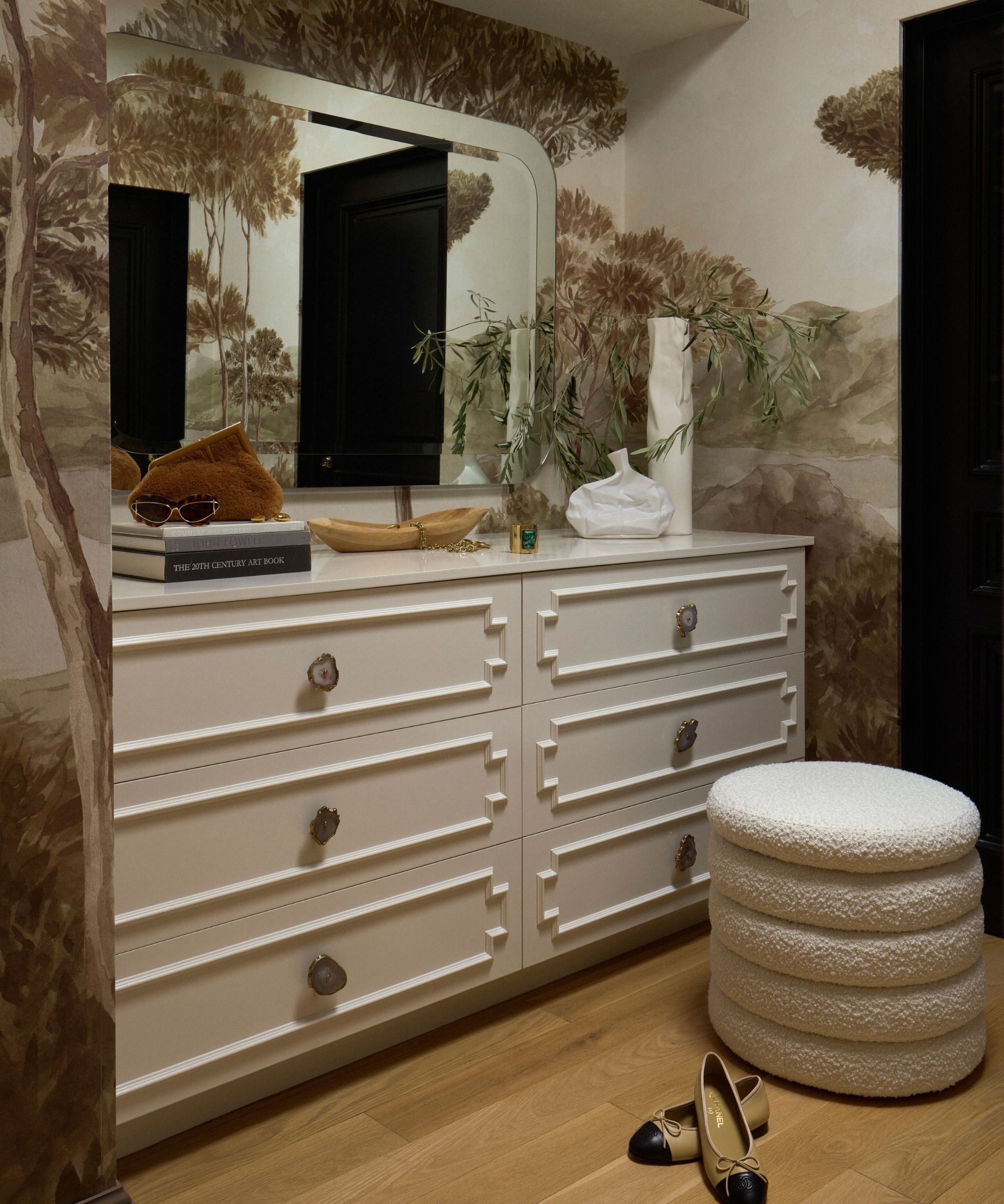 Walk-in closet with white cabinet, mirror on wall above, ornaments and book on top. Wooden floor with round ottoman/seat, mural wallpaper on walls behind
