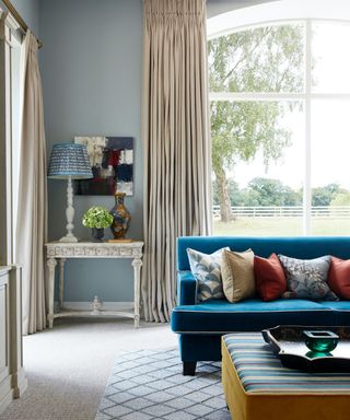 colors that go with light grey, grey living room with cream carpet and drapes, bright blue couch with coloured pillows, stripe foot stool, console table, artwork, lamps, large window with view of garden