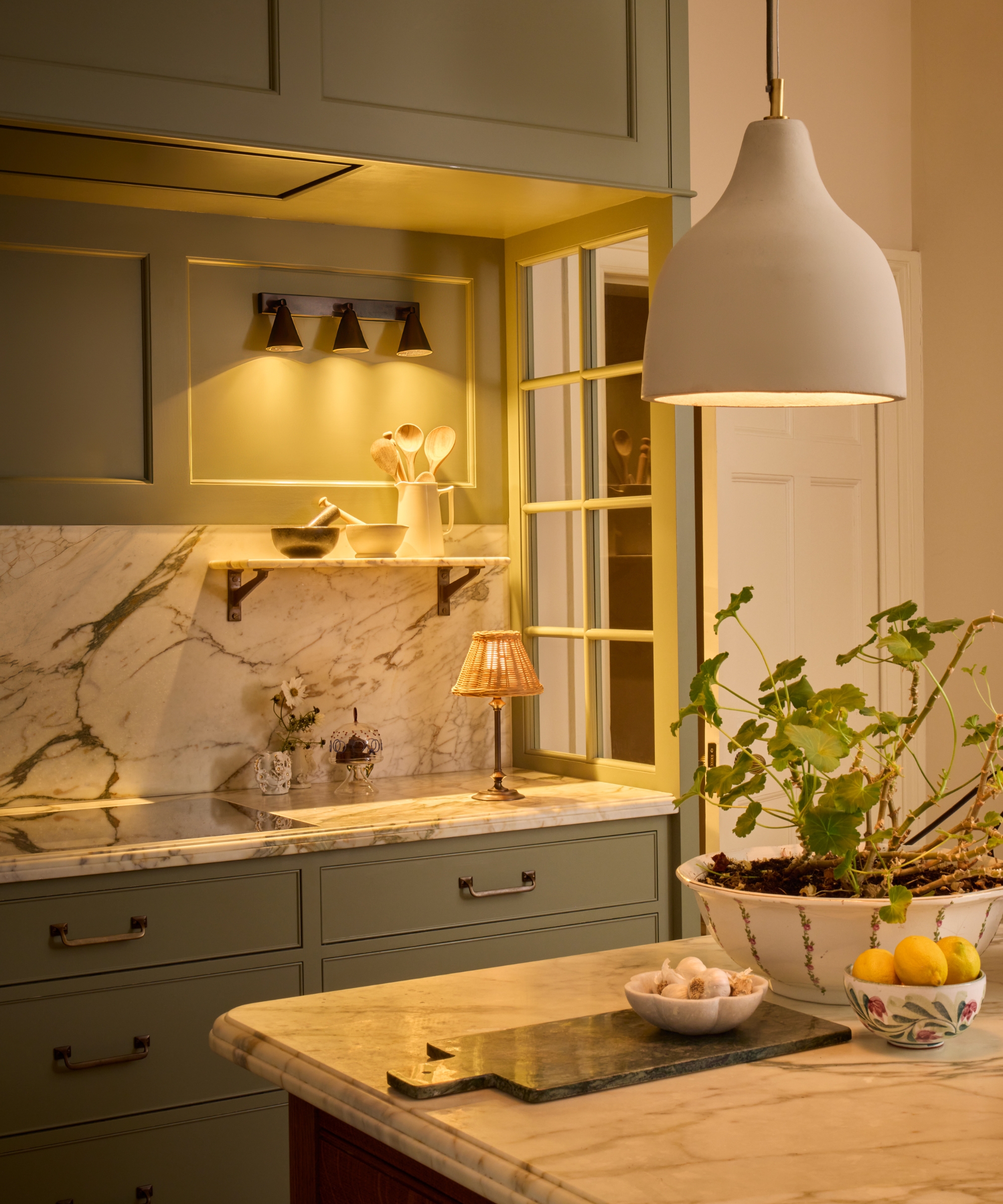 The corner of a kitchen with light green cabinets, a marble countersplash, and lighting with warmer temperature bulbs