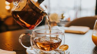 Hand pouring a cup of tea from a stylish transparent teapot into a clear cup.