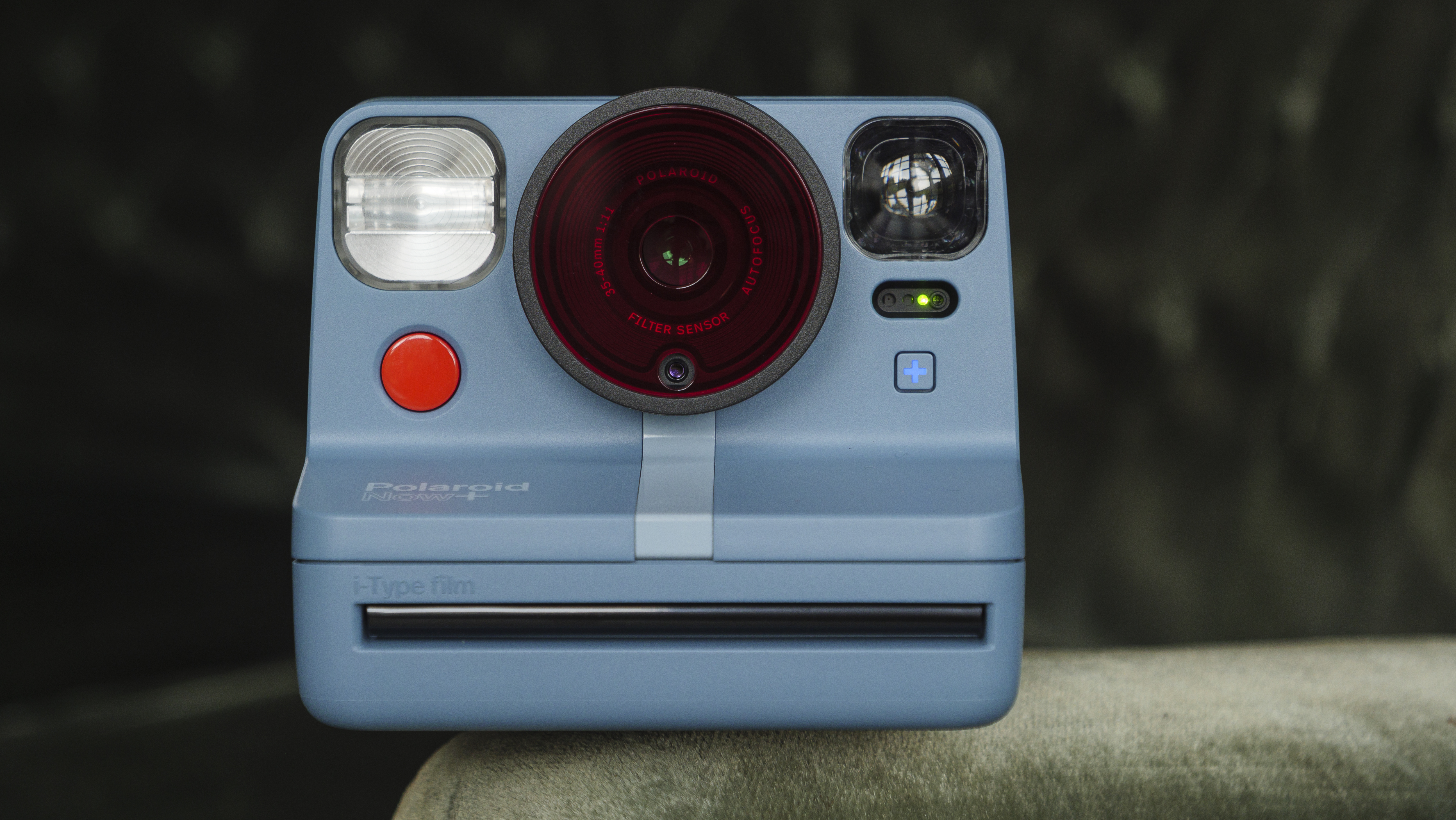 Et blått Polaroid Now Plus-kamera i retrostil står på en sofakant mot en mørk bakgrunn.