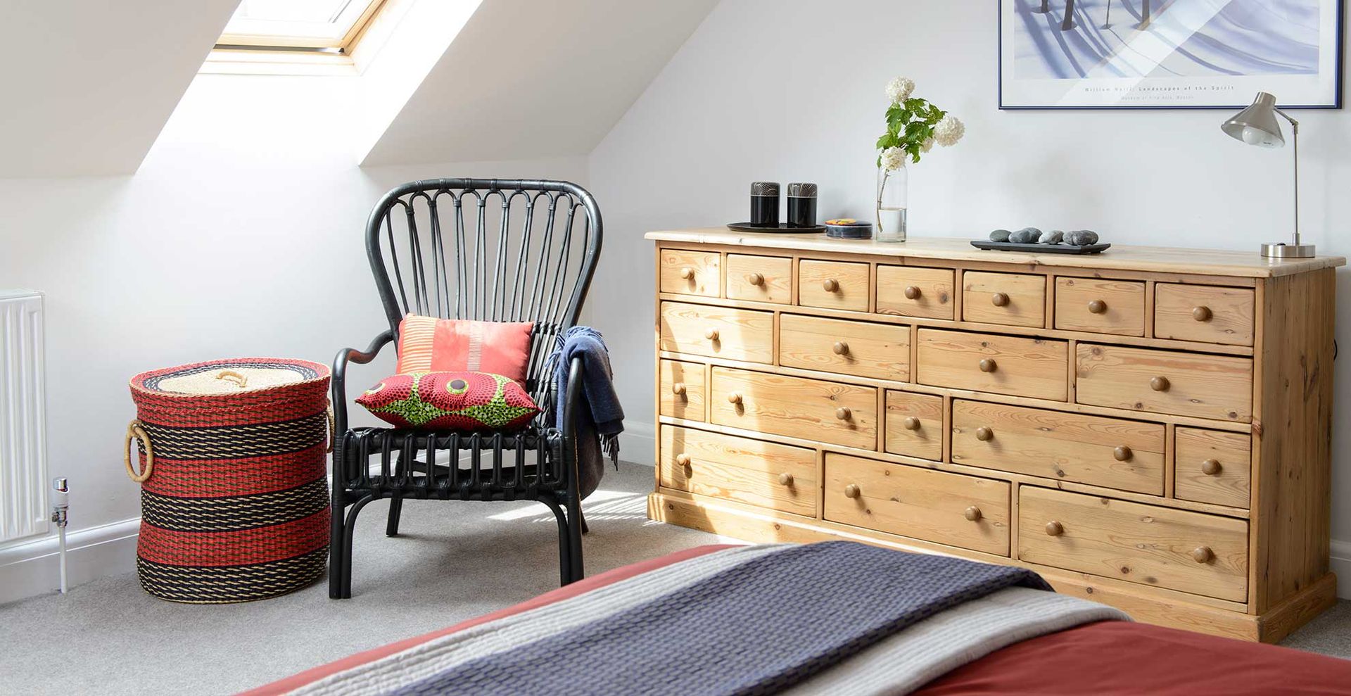 Neutral bedroom with black wicker chair next to a wooden chest of drawers and a laundry basket to show where you should keep a laundry basket