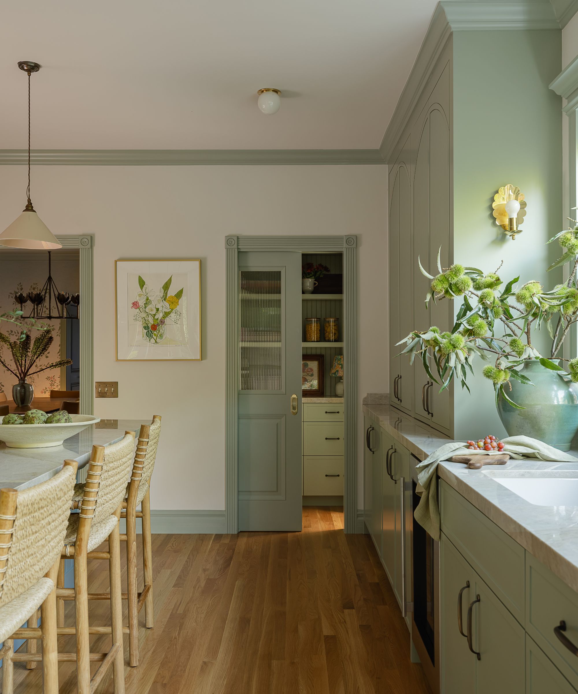 A kitchen view showing a pocket door partially open to a pantry. To the right, tall sage green cupboards reach the ceiling, and a gold scalloped sconce lights a countertop decorated with a large green vase and fresh branches