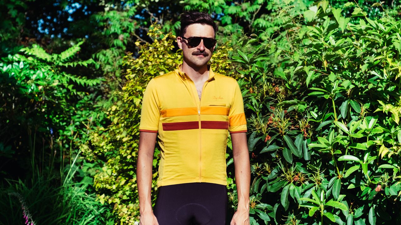 A white man with a moustache and sunglasses wears a yellow jersey