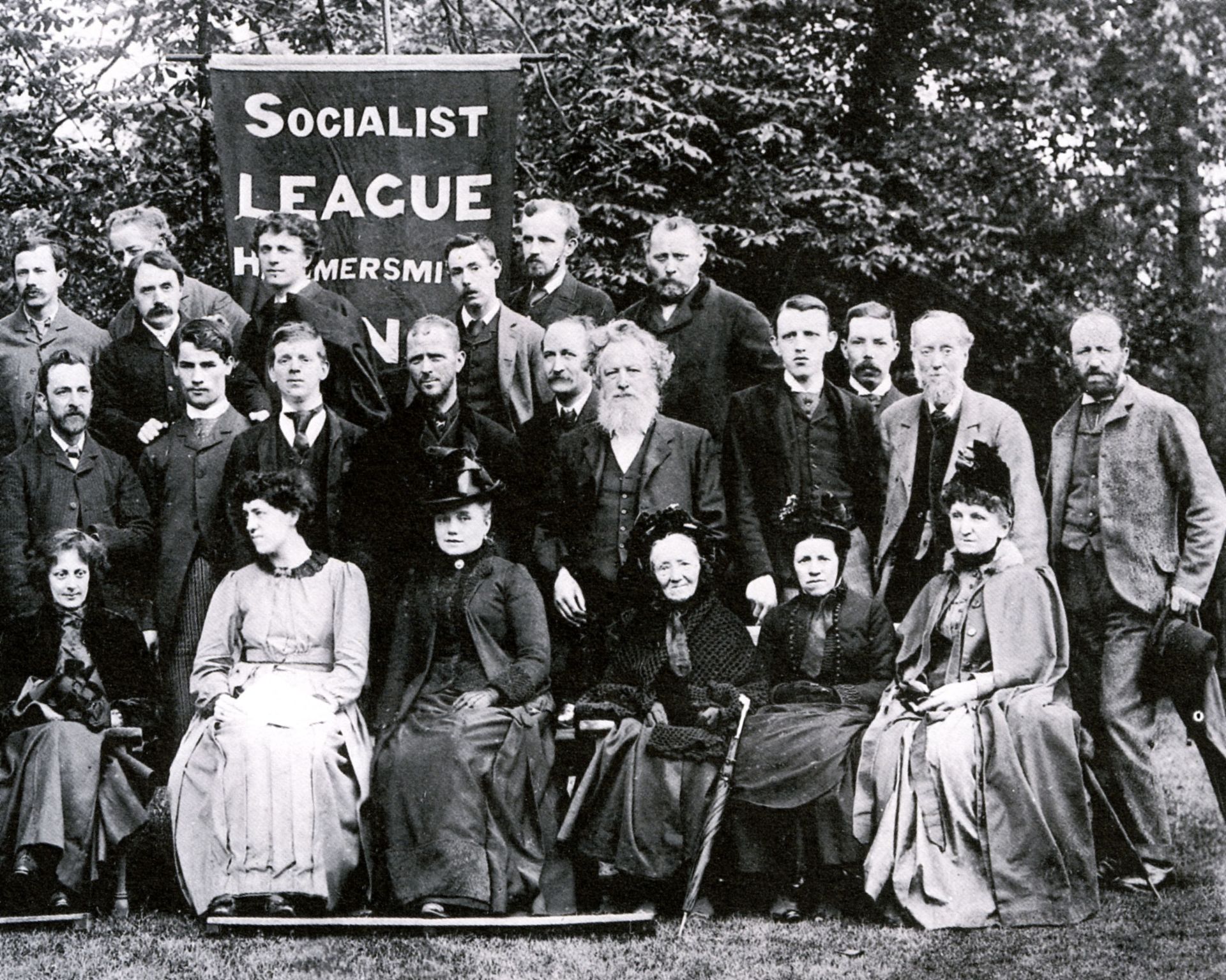 William Morris and other people pose for a photograph in three lines. Someone at the back is holding a banner that reads 'Socialist league Hammersmith'