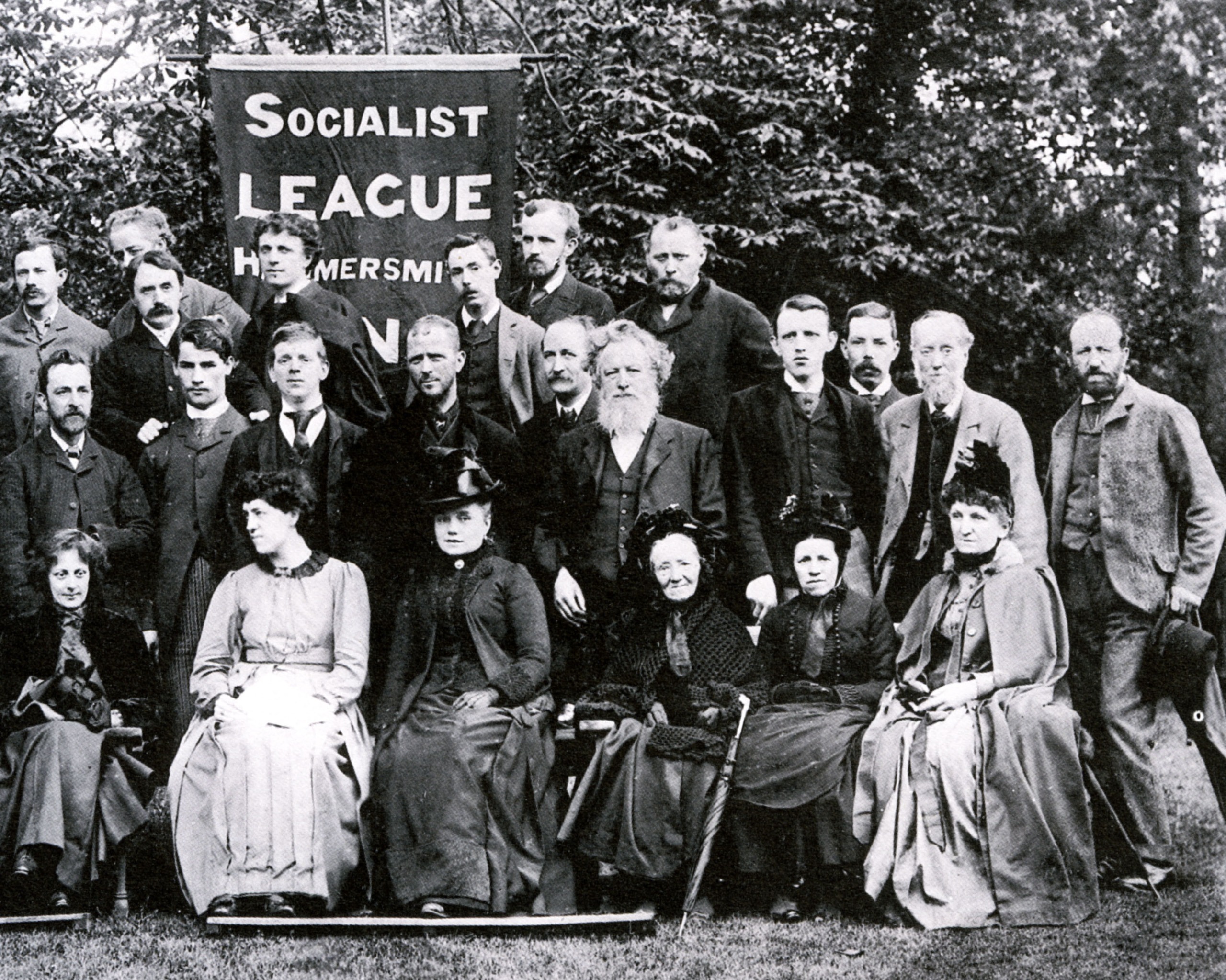 William Morris and other people pose for a photograph in three lines. Someone at the back is holding a banner that reads 'Socialist league Hammersmith'