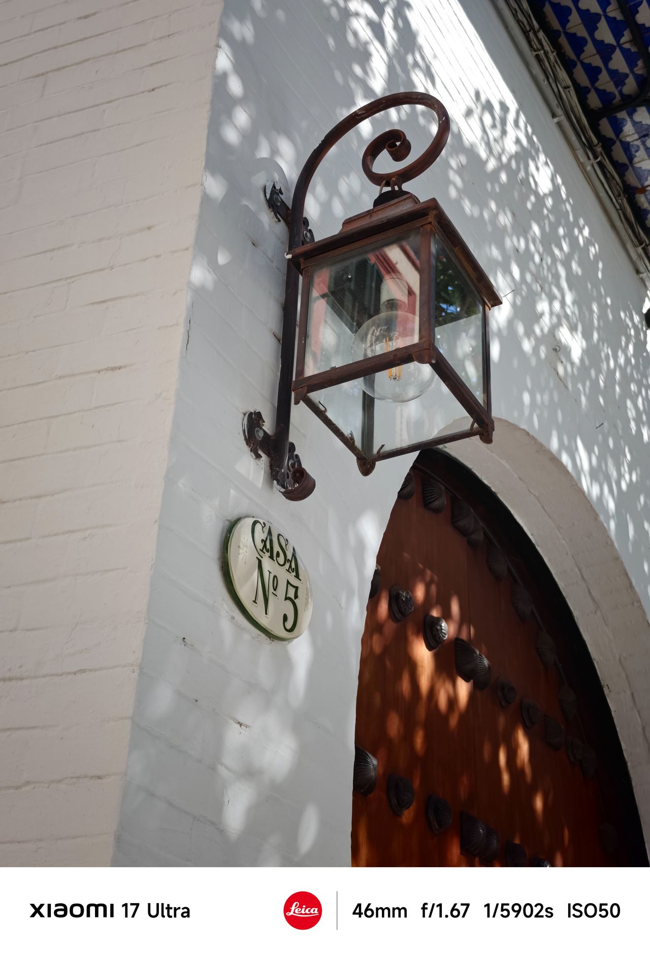 Rustic wall-mounted lantern above a wooden arched door marked &ldquo;Casa N&ordm;5,&rdquo; dappled with sunlight and shadows.