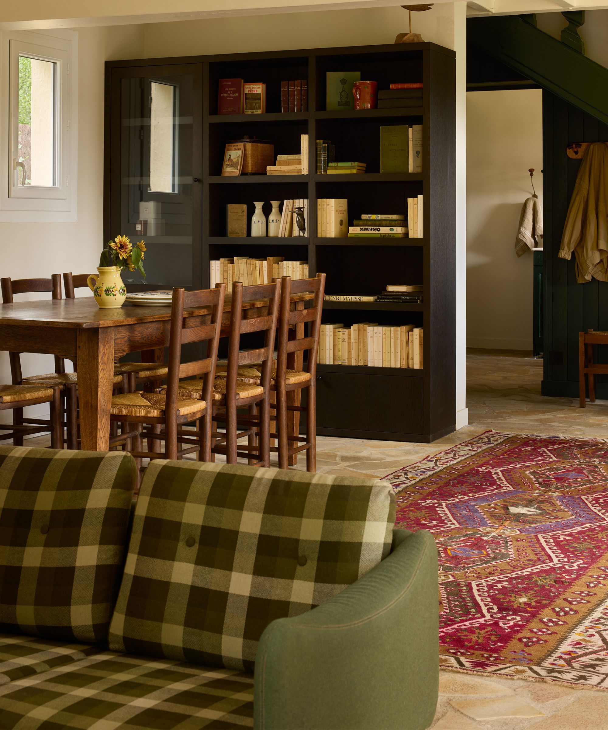 an open plan living dining room with dark timber bookcase, vintage wooden table and chairs, plaid sofa and a red vintage rug