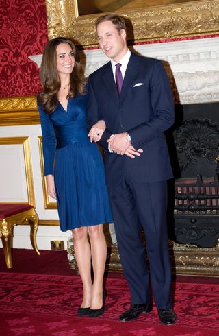 Kate Middleton wearing a blue wrap dress and Prince William wearing a navy suit standing on a red carpet in front of a fireplace for their engagement photo