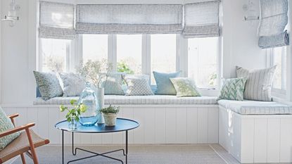 A bright bay window seating area with large windows, sheer grey blinds, patterned cushions, and a circular coffee table with flowers and a large vase, with a wooden rattan chair.