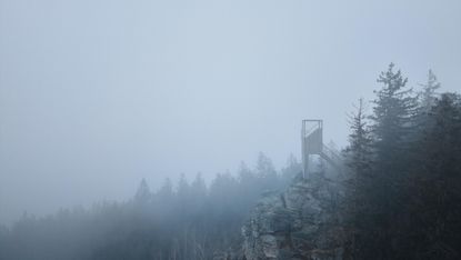 The Miner by Mjölk Architects is a mountain viewpoint, seen here on rocky outcrop, with backdrop of trees, in low cloud