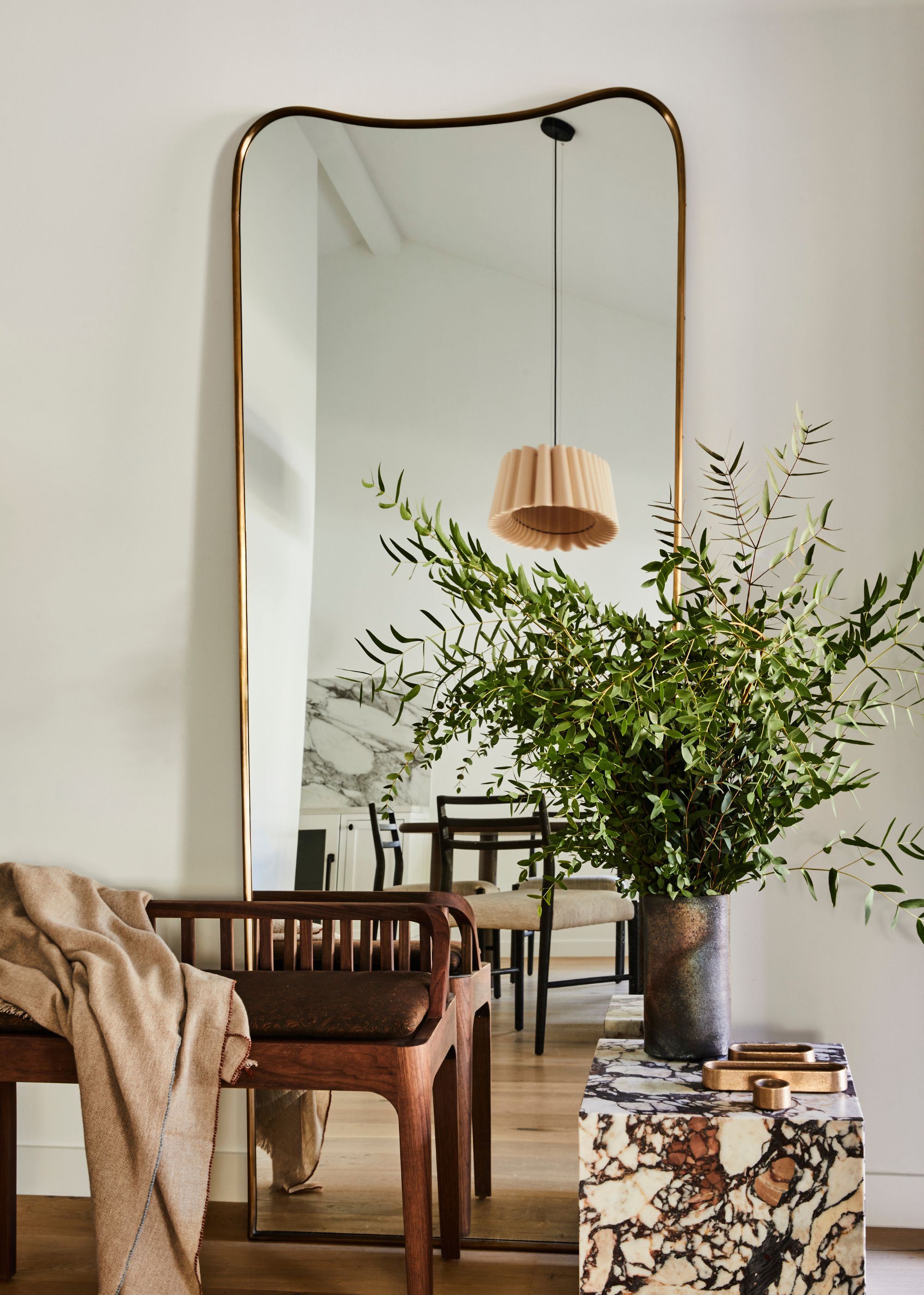 white room with a modern tall mirror against with wall with curved edges, a marble side table with greenery in a vase