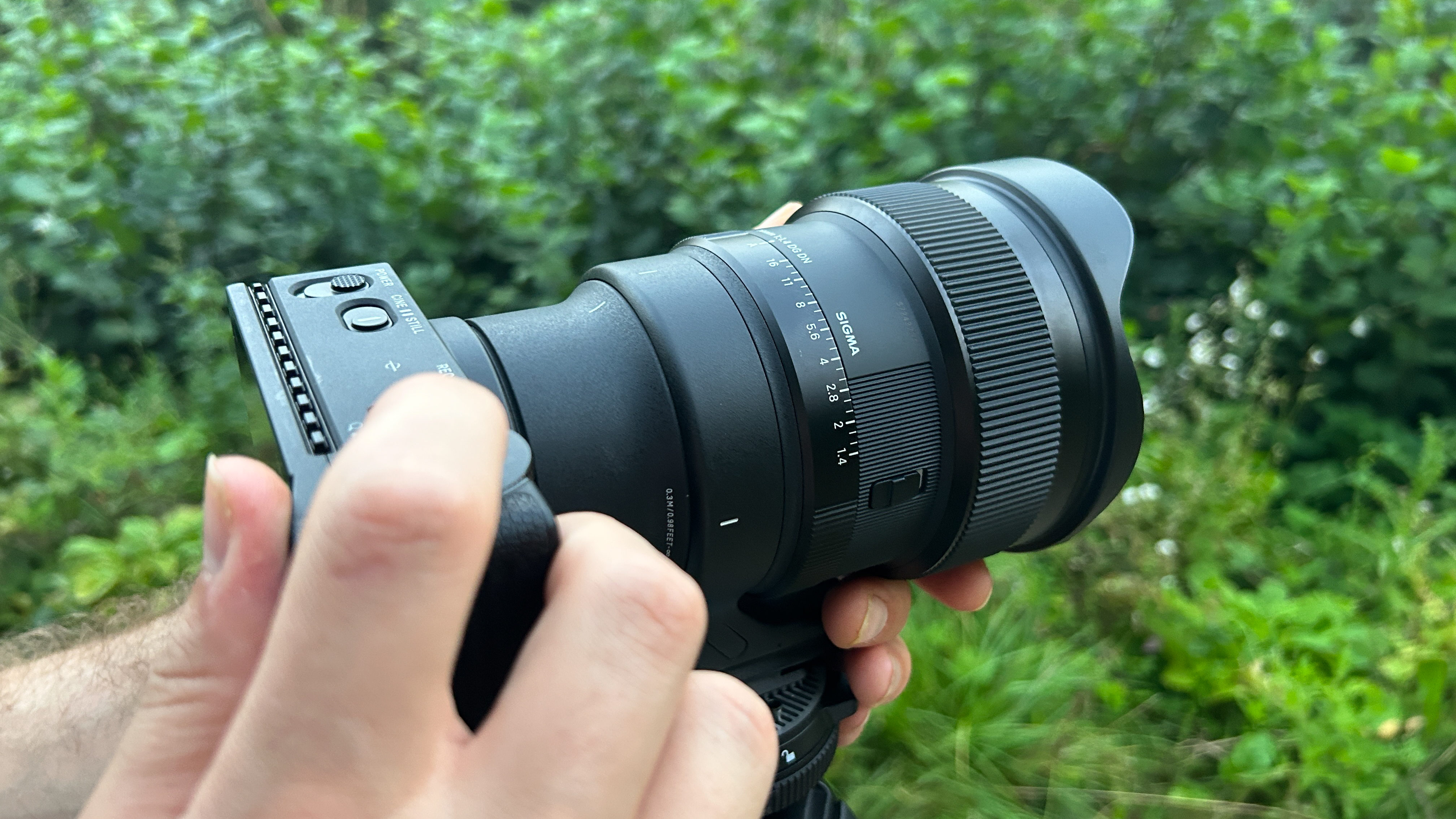 A black camera lens with two ridged adjustment rings is mounted to an out-of-frame tripod and attached to a rectangular black camera. Hands support the item at the mount base of the tripod and on the camera. An index finger rests on the camera trigger button.