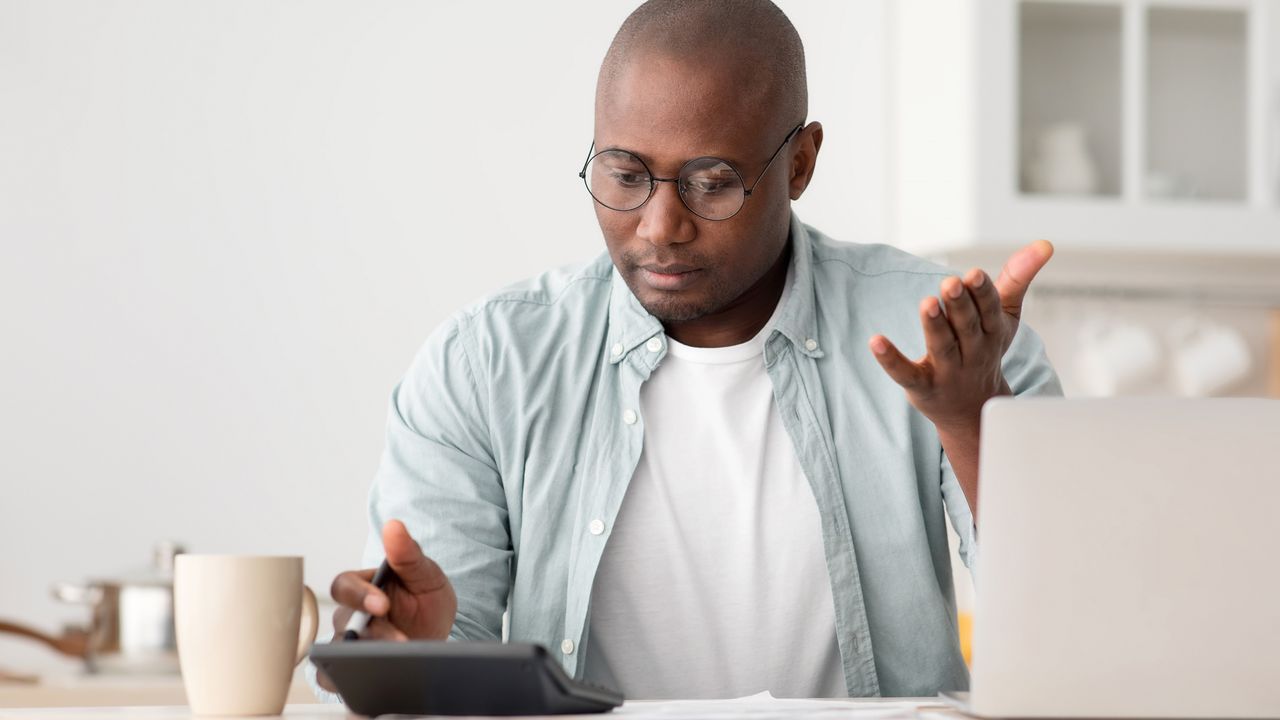 A man looks at his calculator on his desk and makes a &quot;why is that number so high?&quot; gesture.