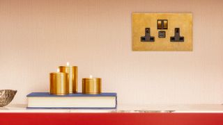 Brass plug socket set against light textured wallpaper with brass candles on book on shelf with red trim
