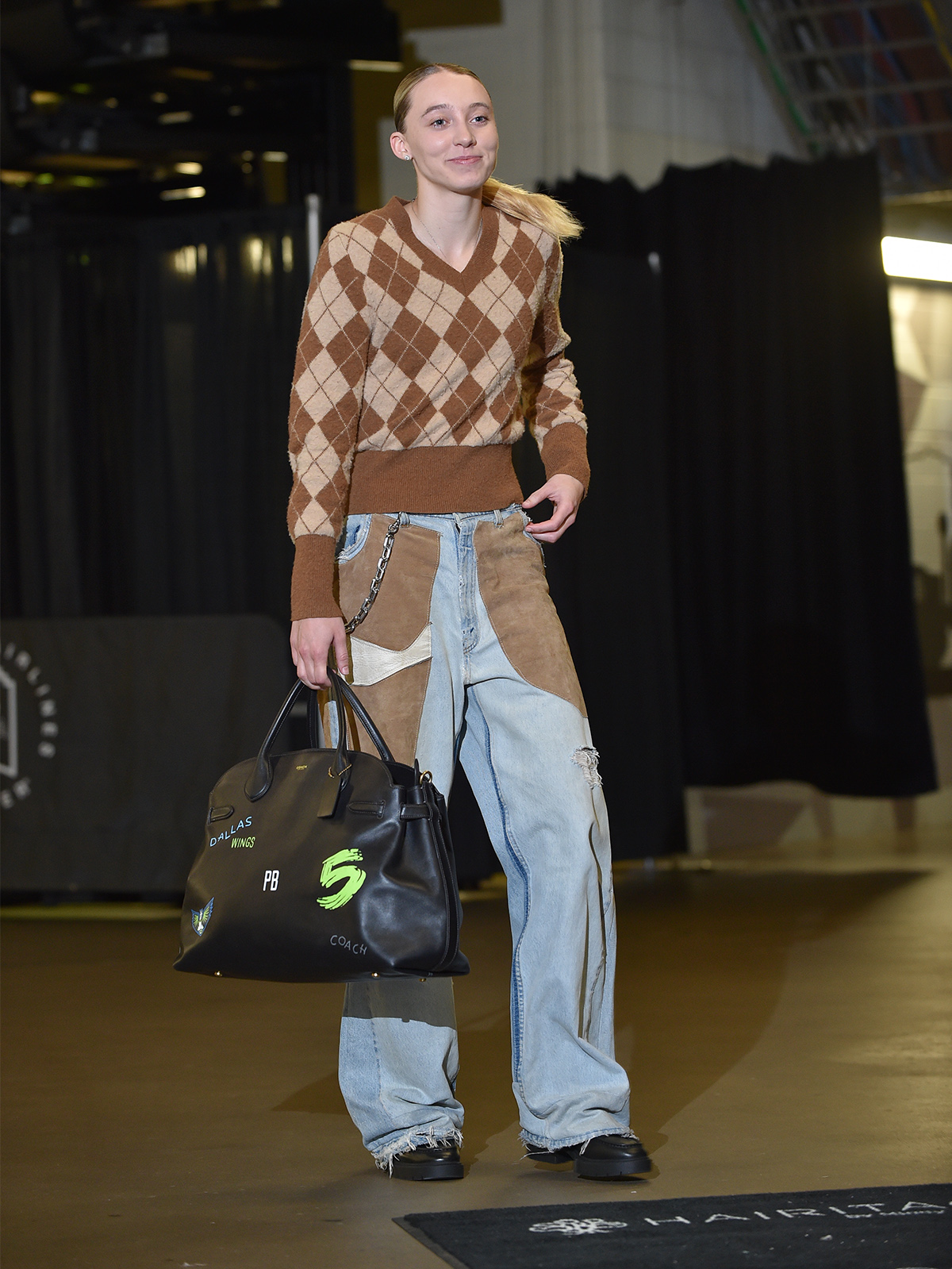 ARLINGTON, TX, August 1, 2025 
 WNBA player Paige Bueckers wears a Coach Fall 2025 shrunken argyle sweater, repurposed suede patchwork jeans, Curb Chain Strap, Leah Loafer, and the Soft Empire Carryall Bag 48 to the Dallas Wings vs. Indiana Fever game.