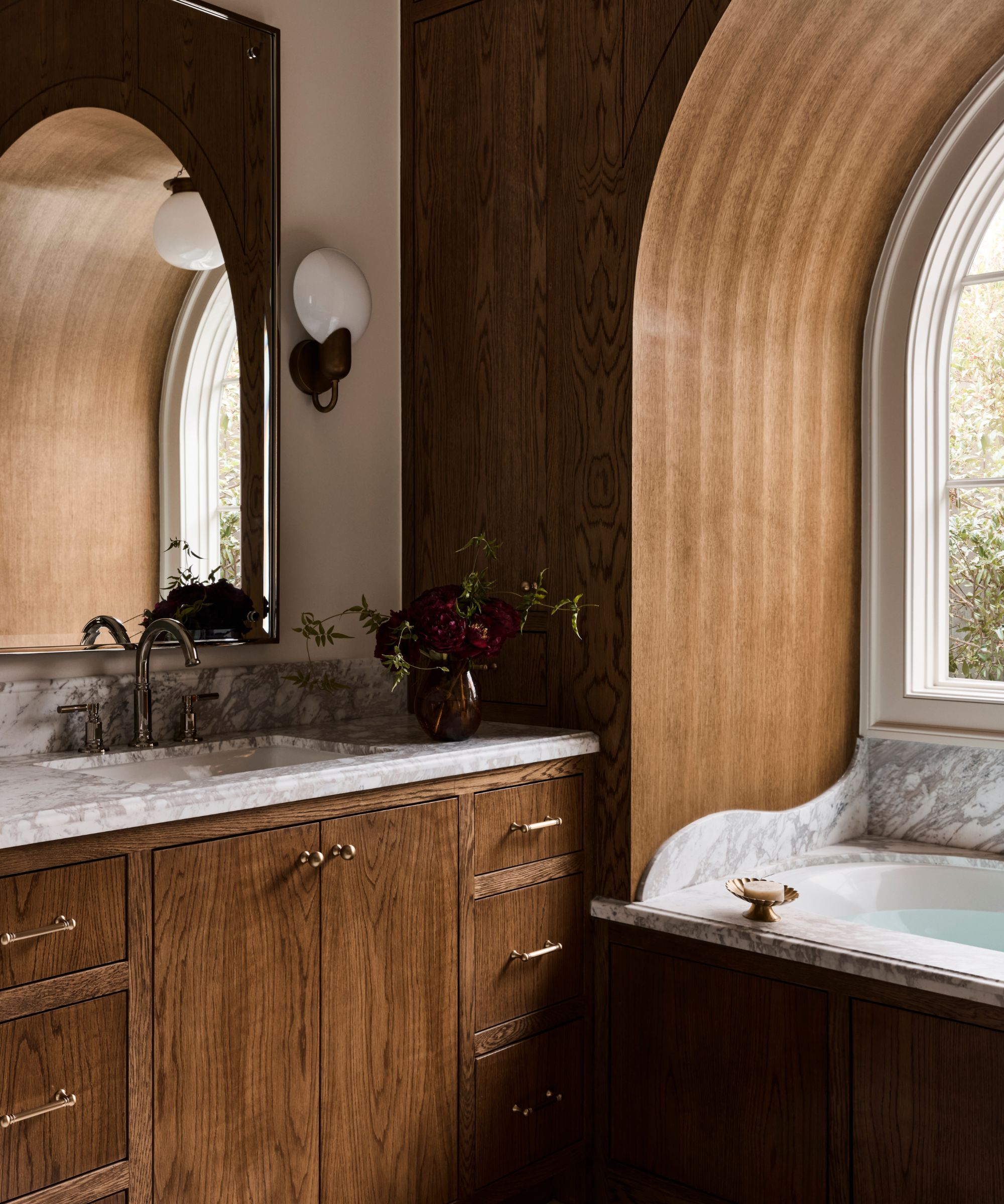Warm bathroom design with rich wood paneling, an arched window nook, white marble countertops, and a built-in bathtub.