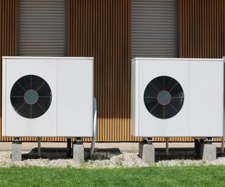 two air source heat pumps stood on ground in front of wooden panels of house exterior