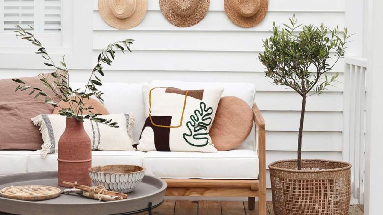 Sofa with cream and brown cushions, coffee table, olive tree in a basket on a wooden verandah deck outside a white wood clad house.