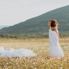 Young cute curly woman in white dress standing near soft linen bed in summer chamomile field. Summer vacations inspiration