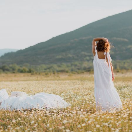 Young cute curly woman in white dress standing near soft linen bed in summer chamomile field. Summer vacations inspiration