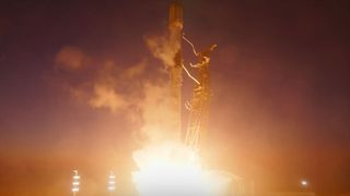 a white and black rocket lifts off into a dusk sky, its bright thrust lighting up the landscape below