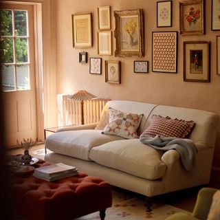 living room with pale plaster pink walls, cream sofa and red buttoned footstool with gallery wall behind